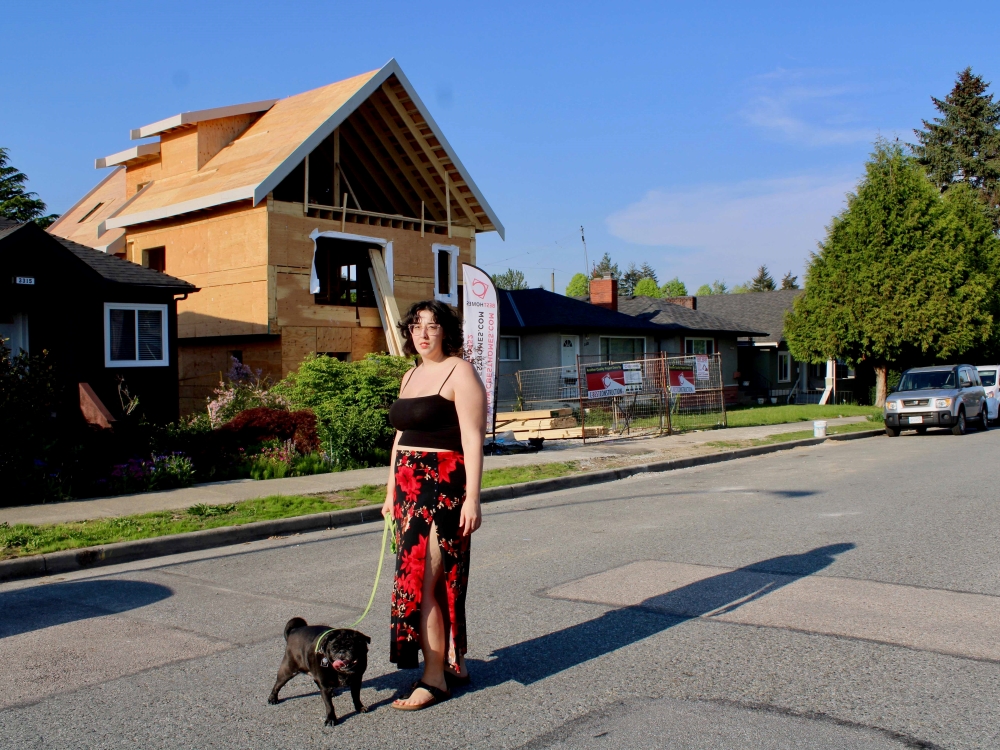 A woman with dark hair wearing a long floral skirt and black tank top stands in front of a house under construction. She holds the leash of a black pug dog.