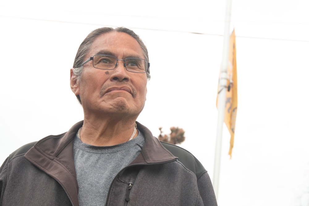 A man wearing glasses and with his hair tied back in a pony tail gazes past the camera. Behind him, an orange flag flies from a pole.