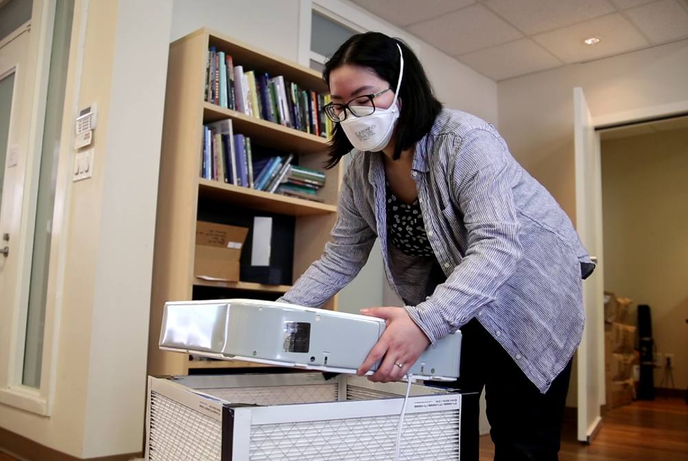 Kimberly Sayson bends slightly to place a fan on top of the filter frame. She is wearing a blue striped button-down shirt.