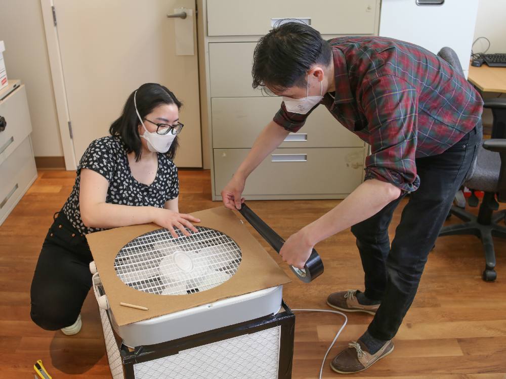Kimberly Sayson, left, has shoulder-length black hair and glasses. She is wearing a black and white floral shirt and crouches to hold a piece of cardboard with a wide circle cutout in place while Jeffery Chong, right, secures it with black duct tape. He is standing and bending slightly, wearing a red plaid shirt and black jeans. Both are wearing white masks.