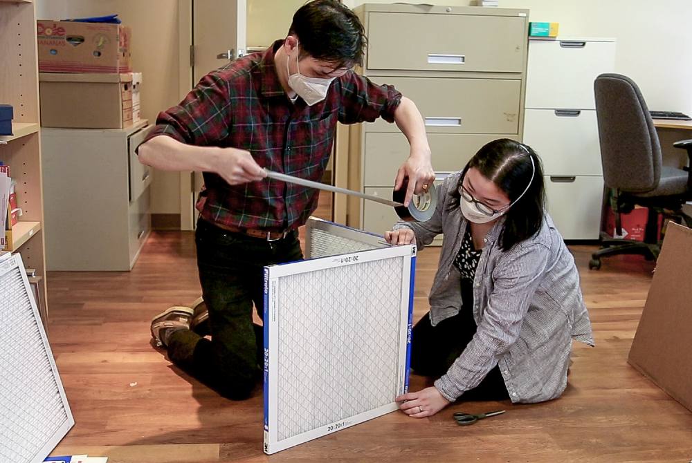 On the left, Jeffery Chong kneels over two white air filters while rolling out a piece of duct tape to tape them together. On the right, Kimberly Sayson holds the filters in place.