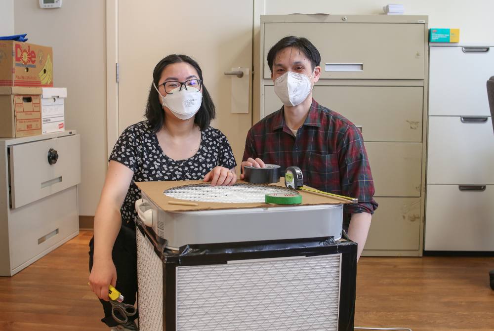  Kimberly Sayson, left, and Jeffery Chong, right, crouch behind a white Corsi-Rosenthal box outfitted with black duct tape and brown cardboard in an office setting with brown wood floors. They have black hair, are dressed casually and are wearing white masks. Their eyes indicate they are smiling.