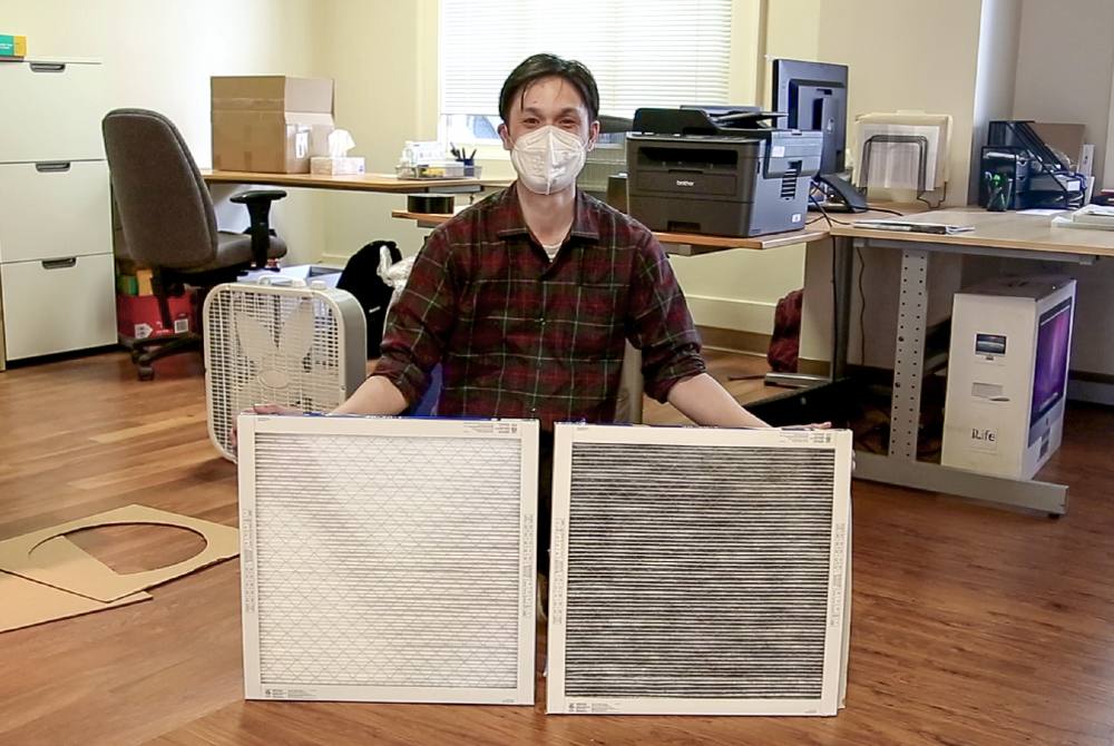 Jeffery Chong kneels behind two white square air filters, holding them side by side in each hand. He is wearing a red plaid shirt and white mask. On the left, a new clean filter is white and untarnished. On the right, an older used filter is dark and grey.