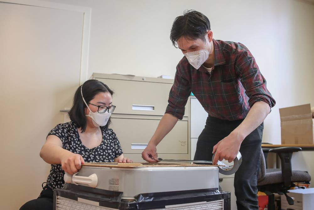 Kimberly Sayson kneels behind a Corsi-Rosenthal box on the left while Jeffery Chong, right, seals the edges generously with duct tape.