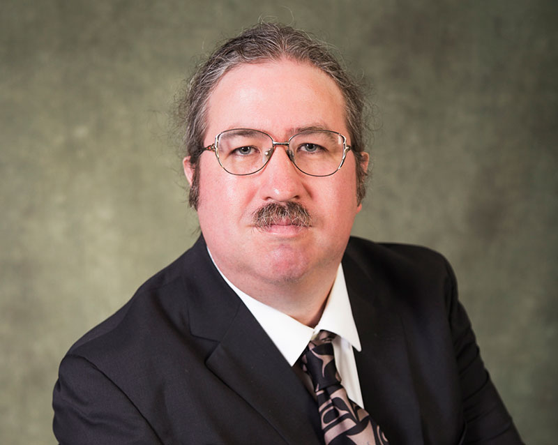 A man wearing a suit and tie and glasses, with a moustache, and his hair pulled back, looks steadily at the camera.