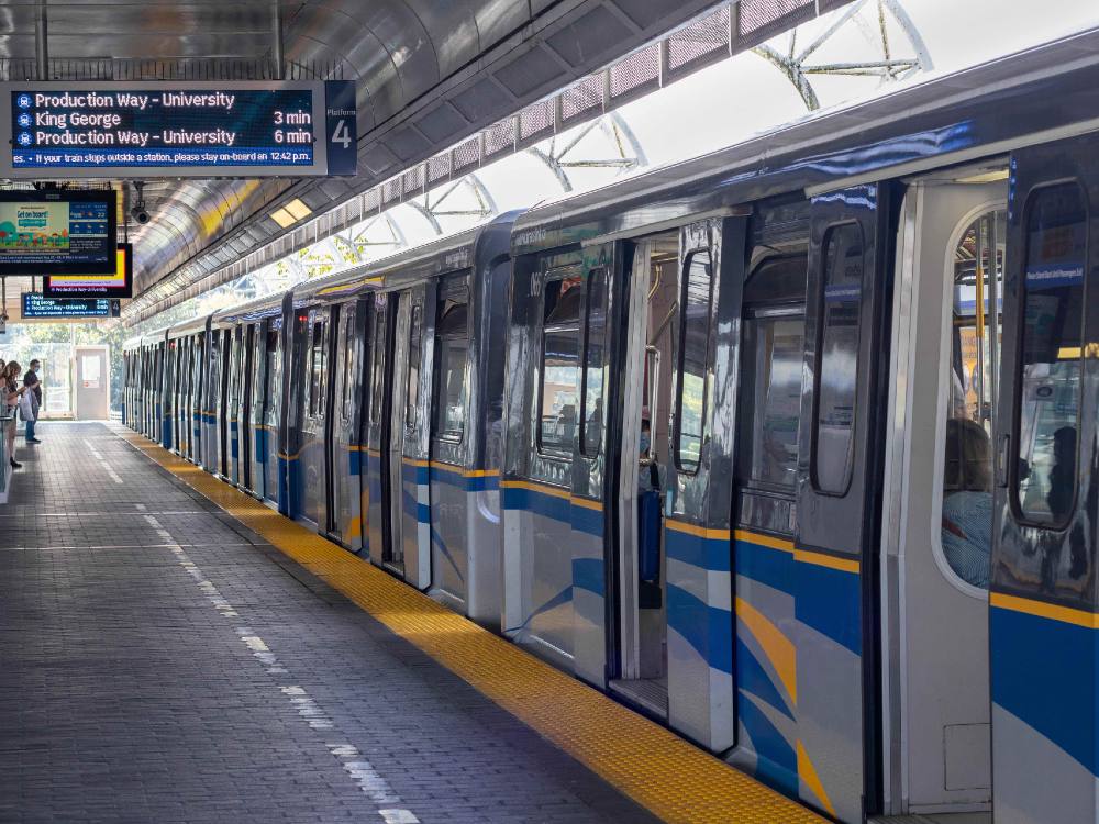 A photo of a SkyTrain station. A train is on the right, doors open and paint shining. Some passengers are standing near the end of the platform.