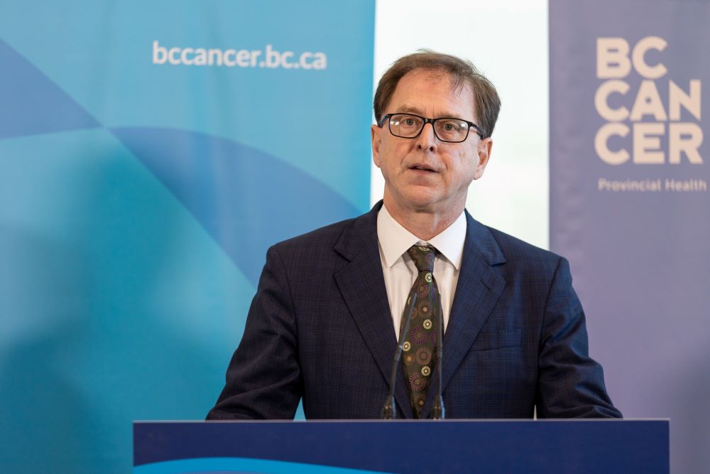 Adrian Dix, wearing a suit, stands behind podium. Behind him a backdrop says bc.cancer.bc.ca.