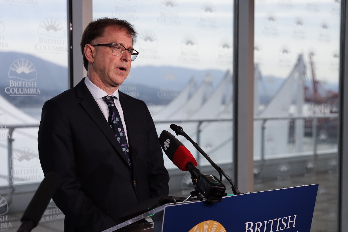 Adrian Dix, in a dark suit and tie, speaks from behind a podium. Canada Place is visible through the windows behind him. 