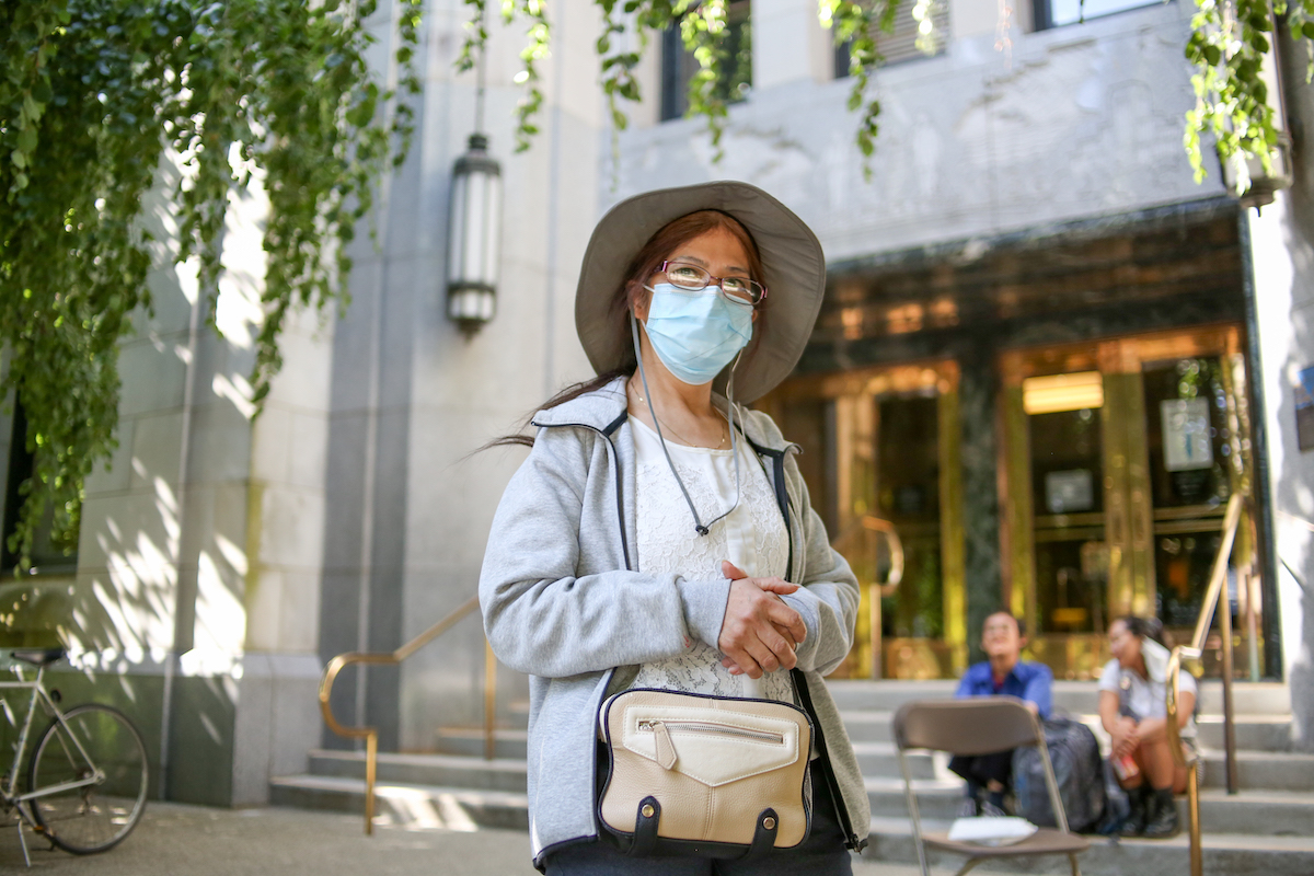 A Chinese woman in her early 60s. She’s wearing a mask and a sun hat. She’s standing in front of the steps of Vancouver City Hall.