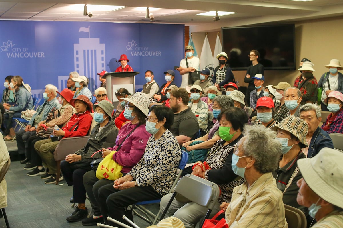 Chinese seniors in colourful hats and jackets pack an office meeting room with a backdrop of a drawing of Vancouver City Hall.