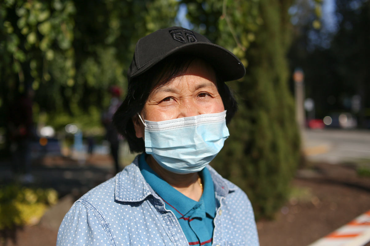 A Chinese woman in her early 60s. She’s wearing a mask. She is wearing a white-spotted denim shirt over a teal polo.
