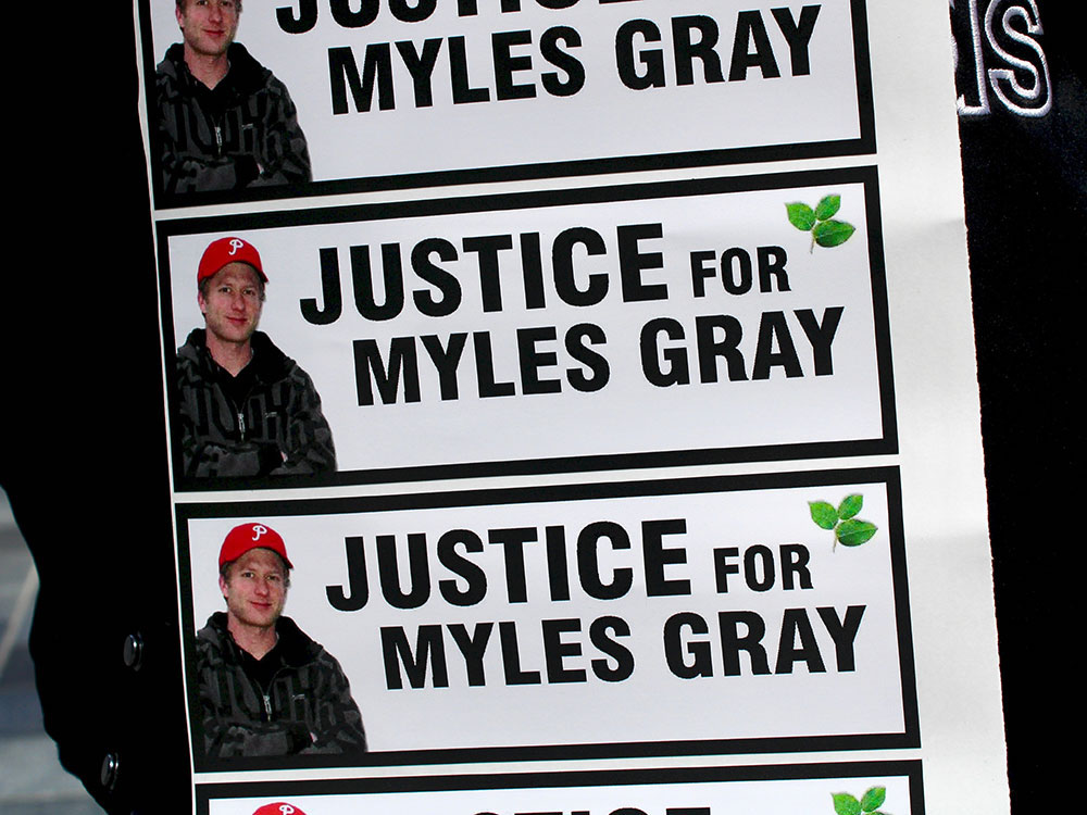 A person holds a sheet of stickers that read “Justice for Myles Gray” in large black letters. The stickers include a photo of a man wearing a red ball cap.