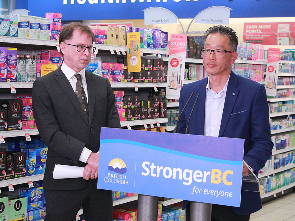 Health Minister Adrian Dix looks on as Chris Chiew stands behind a podium set up. A shelf of drug store products is behind them.