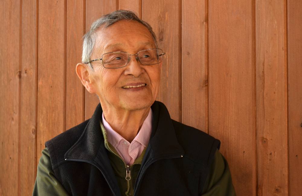 An older man with grey hair and glasses smiles, looking off to his left. He's wearing a pink shirt, a green jacket and a black fleece vest underneath, with a background of wooden panels.