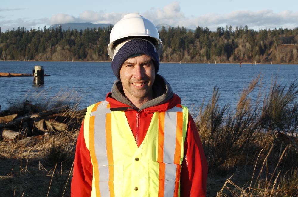 A man wearing a toque underneath his white construction hat and a reflective safety vest smiles with the background of the Fraser River’s north arm behind him.