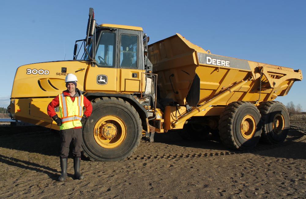 A man in an orange jacket, a reflective safety vest, a white helmet and rubber boots stands beside a large Deere truck with tires that are nearly his height.