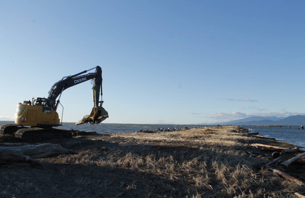 A Deere excavator stands on a narrow piece of grassland besieged by water on both sides and stretches out to the horizon of a blue sky.