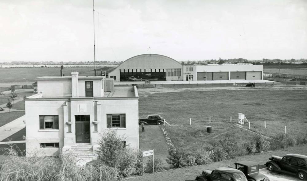 Two structures, one stocky square building and a hangar with a curved, are surrounded by fields and cars that dot the property.