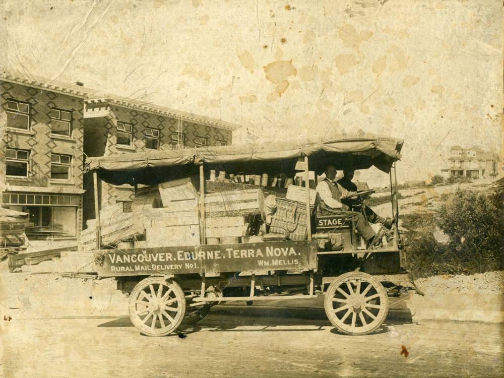 Two men look at the camera from the front seat of a delivery truck. The truck's canvas walls are rolled up and you can see the piles of baskets, boxes and crates heaped in the back. In the background a three-storey building is under construction.