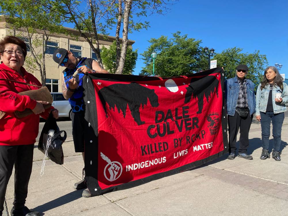 A red and black banner several feet high with the words “Dale Culver killed by RCMP Indigenous lives matter” is held up by two people on either side.