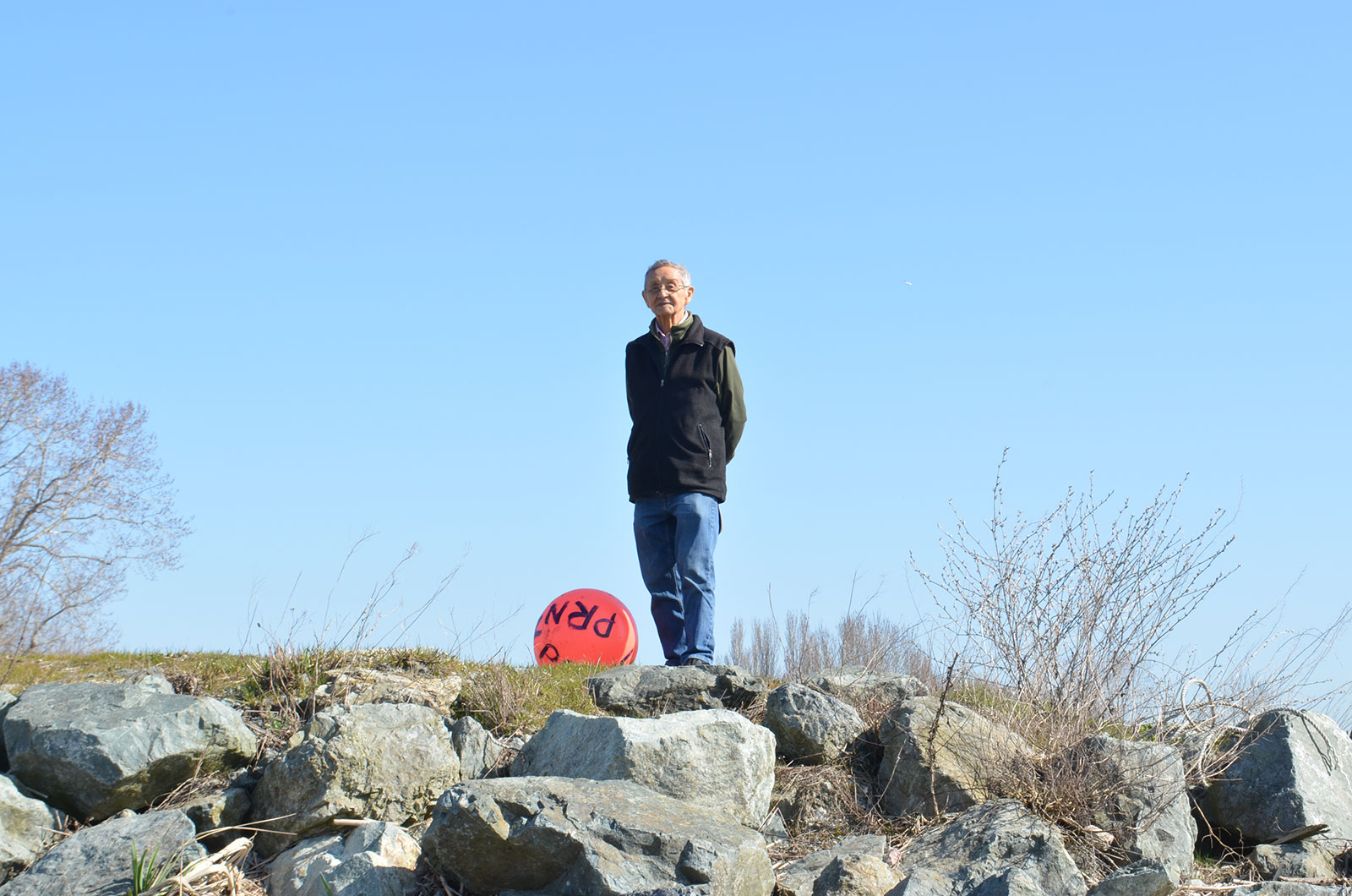 An older man in a black vest and jeans stands on a hill surrounded by rocks, brush and a blue sky.