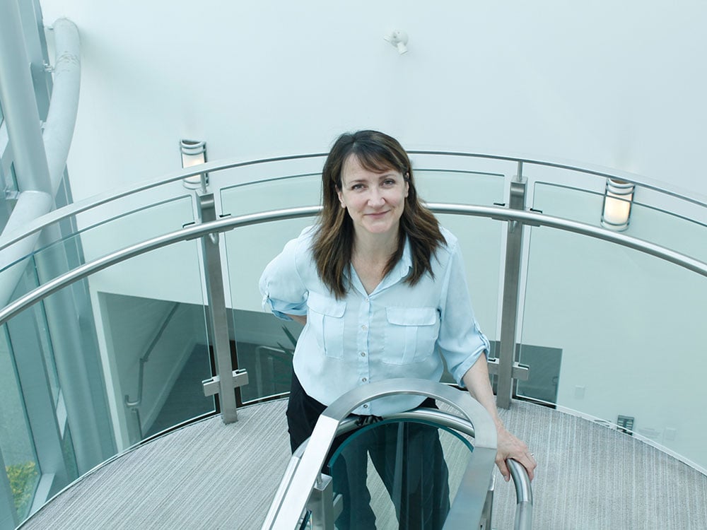 A white woman with brown shoulder-length hair stands in a modern, rounded stair landing looking up at the camera.
