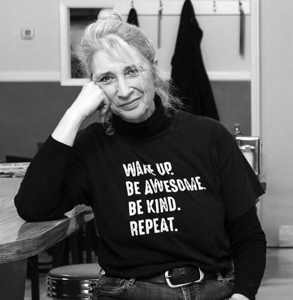 A black and white photo of a woman with her chin leaning on her hand. Her T-shirt reads, “Wake up. Be Awesome. Be Kind. Repeat.”