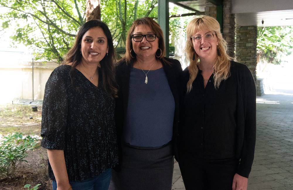 Three women pose for a photo, with their arms around each other, in a courtyard.