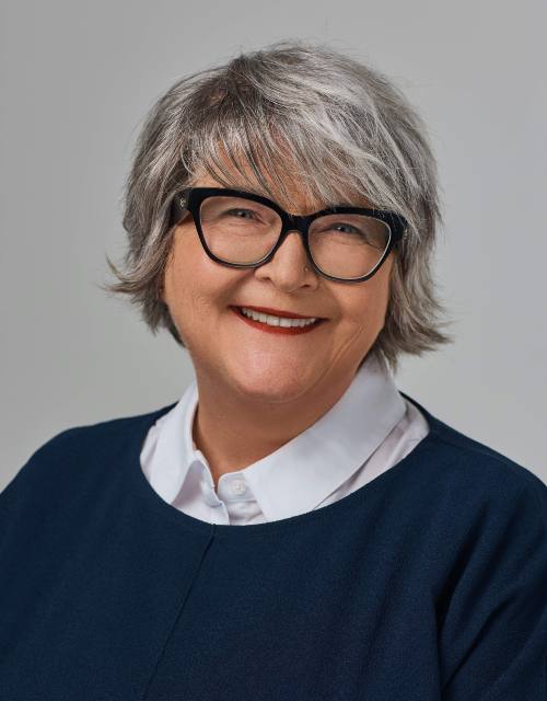 A woman with a salt-and-pepper bob and black-rimmed glasses smiles at the camera.