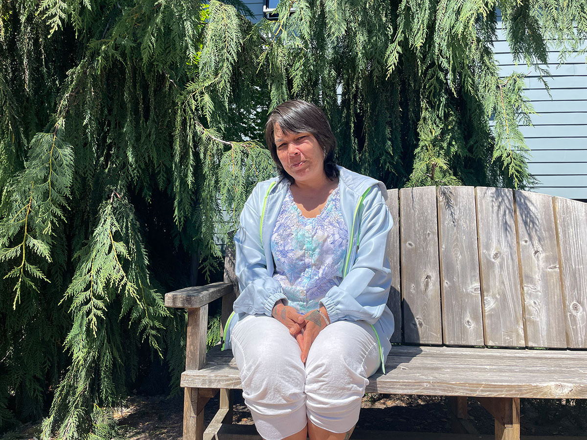 A woman with shoulder-length dark brown hair sits on a wooden bench, looking directly and evenly at the camera.