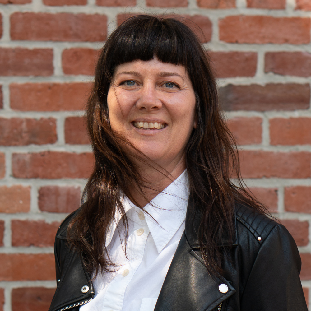 A woman with long, dark hair and bangs, wearing a leather jacket and a white button-up shirt, smiles at the camera.