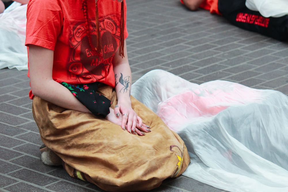 A child lies covered in a white shroud while a woman in an orange “Every Child Matters” T-shirt kneels next to him with folded hands.