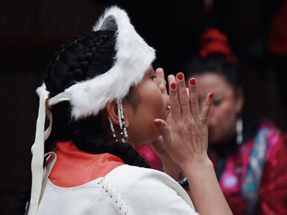 An Indigenous woman holds her hands on either side of her mouth as she closes her eyes during a dance performance. She is wearing red under white. Her dark braided hair is adorned with a white fur beaded headpiece.