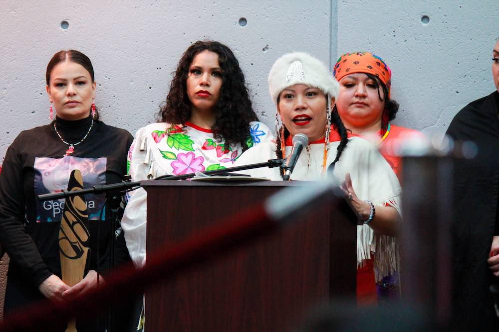 Lorelei Williams is dressed in white and speaks into a microphone. Three women stand behind and alongside her. From left, the women are wearing black, white with a floral pattern and orange.