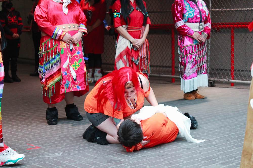 A dancer lays a child covered in a white shroud onto a tiled concrete walkway. Dancers stand around them in a semi-circle.