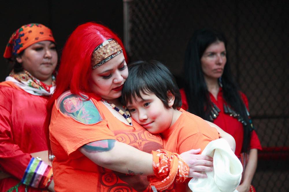 A boy rests on the chest of his mother, who is giving him a hug. They are both wearing orange shirts. Two people stand behind them in the background.