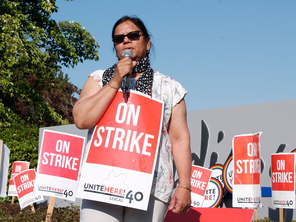 Pardeep Thandi speaks into a microphone at a protest, wearing a black and white scarf and light shirt. Behind are a number of red signs that say, “On Strike.”