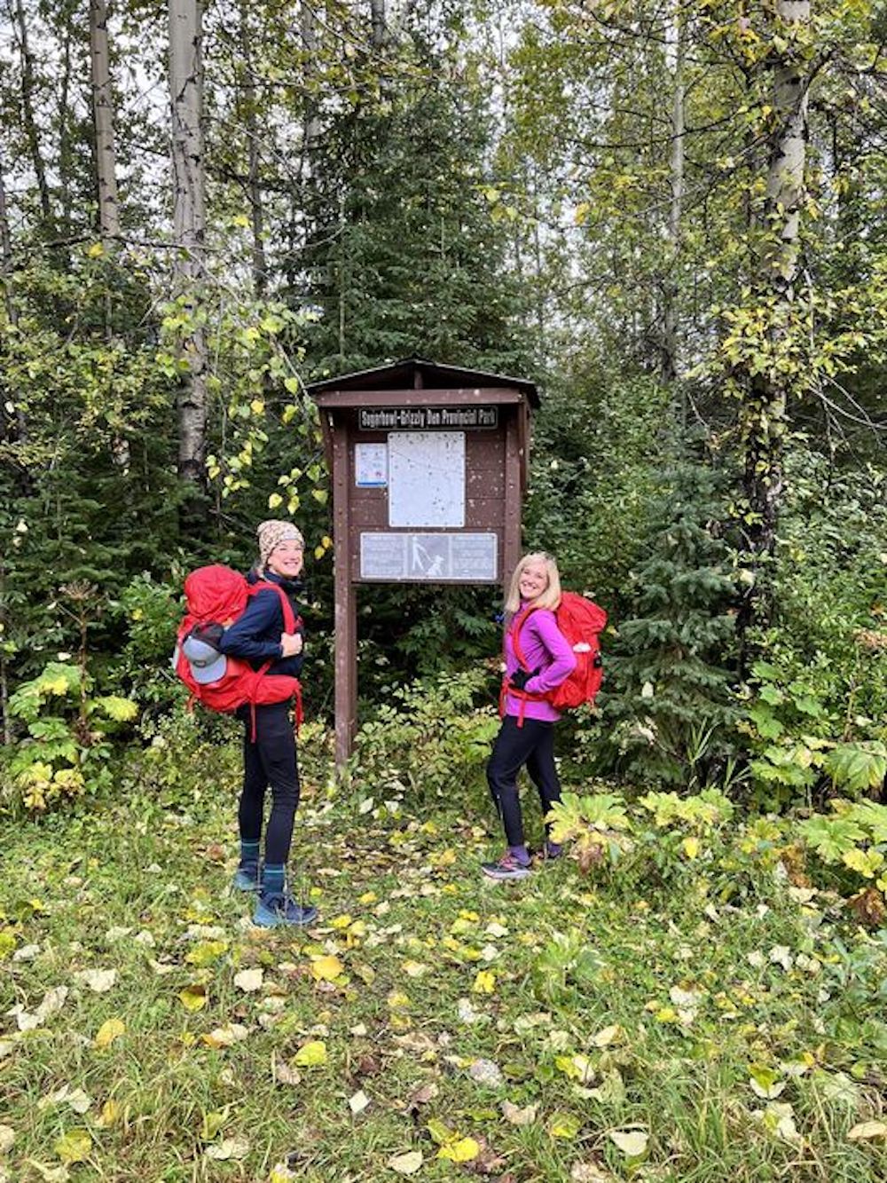 Two women wearing hiking clothes and red backpacks stand at the edge of a forested area, in front of a park sign that reads ‘Sugarbowl-Grizzly Den Park.’
