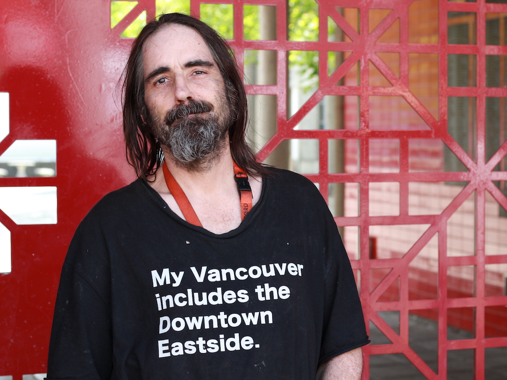 A man with shoulder-length salt-and-pepper hair and a beard looks at the camera. He’s wearing a black T-shirt that reads, ‘My Vancouver includes the Downtown Eastside.’