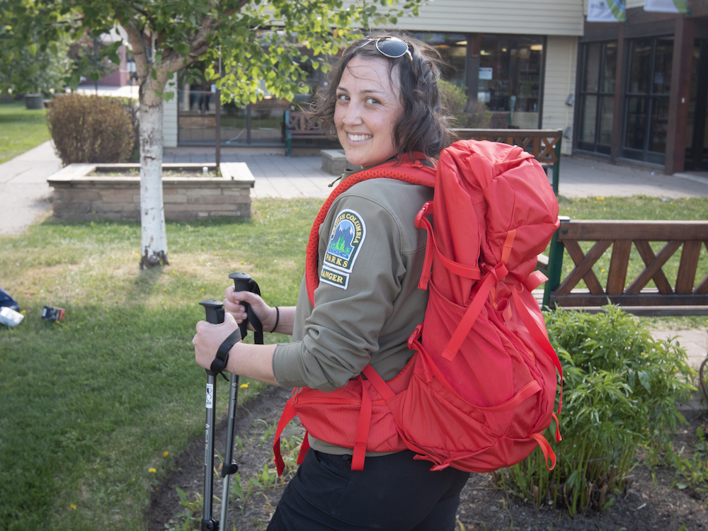 A woman with shoulder-length brown hair and wearing a BC Parks uniform smiles at the camera while wearing a bright red backpack.