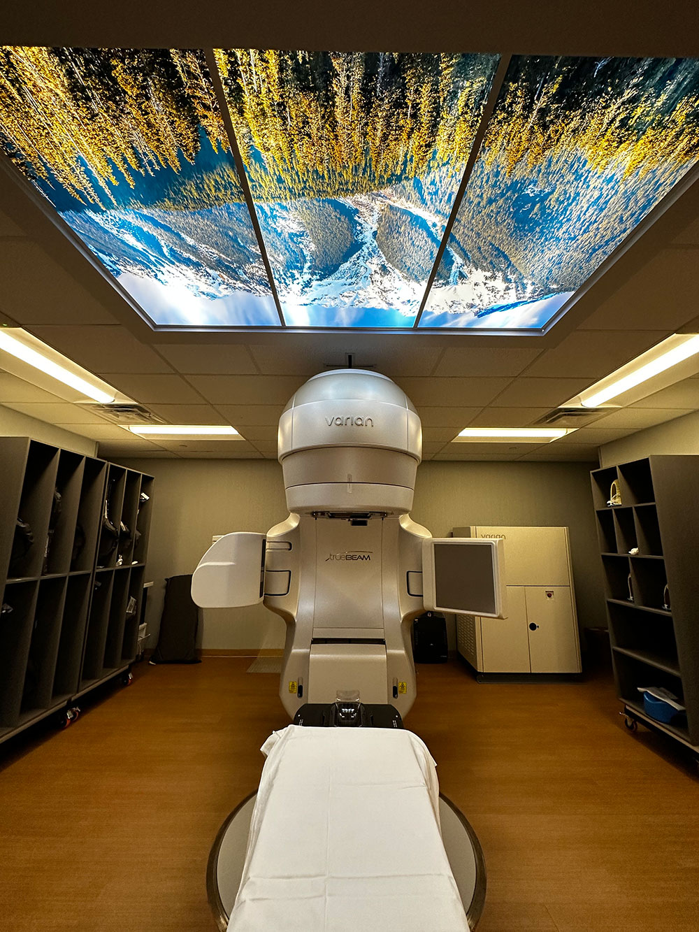 A large digital poster of a sunny mountain range adorns the ceiling of a cancer treatment room featuring a large beige medical apparatus at the North Cascade Cancer Treatment Center in Bellingham, Washington.
