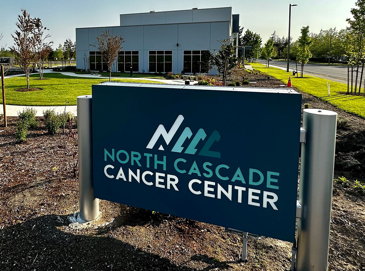 A blue sign with a digital graphic of a mountain range displays the name of the North Cascade Cancer Center in Bellingham, Washington, on a sunny day. Behind it is a short, non-descript light blue building.