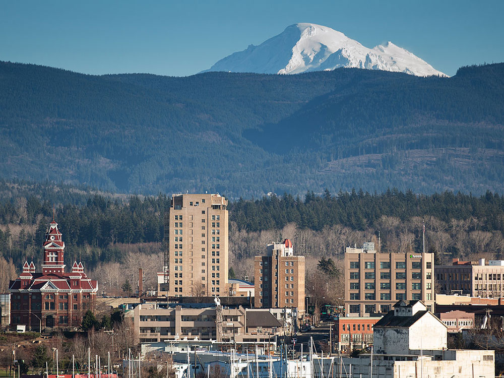A snowcapped peak rises in the background of building tops of a mid-sized city.  