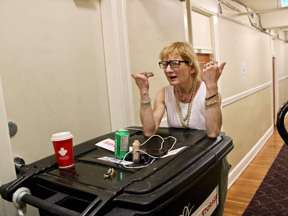 Lisa Welsh, wearing a sleeveless white shirt and bracelets, leans on a tall black plastic bin in a hallway with beige walls and patterned carpet.