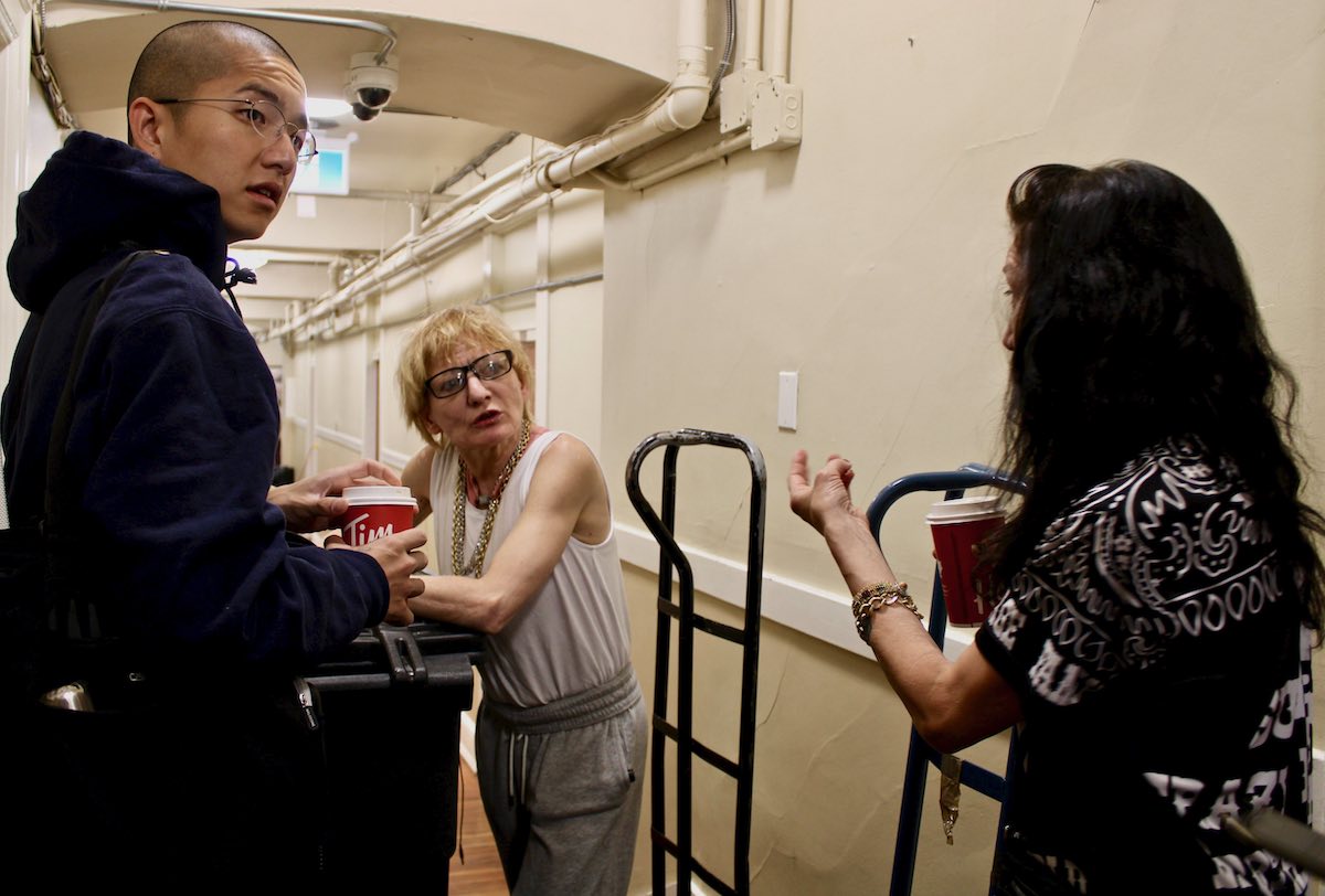 Vince Tao, in a dark hoodie, Lisa Welsh, leaning on a tall black plastic bin, and Wendy Gaspard talk in a hallway.