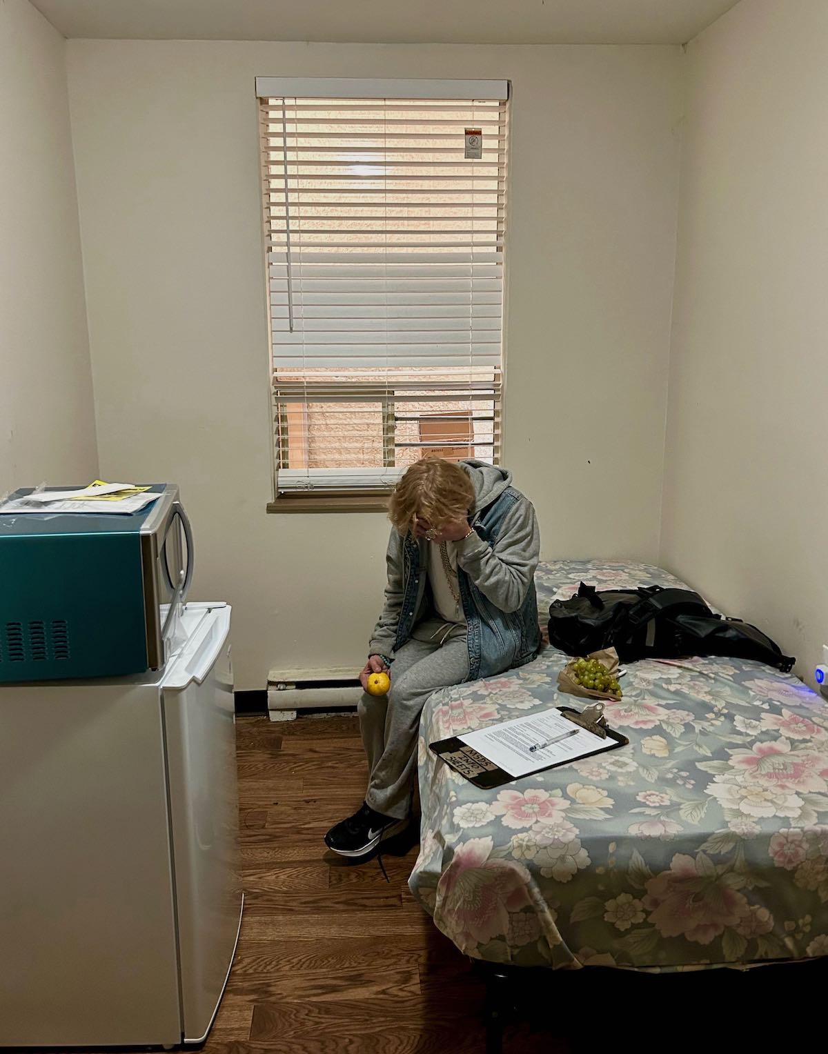 Lisa Welsh sits on a bed in a small room with a microwave and compact refrigerator.