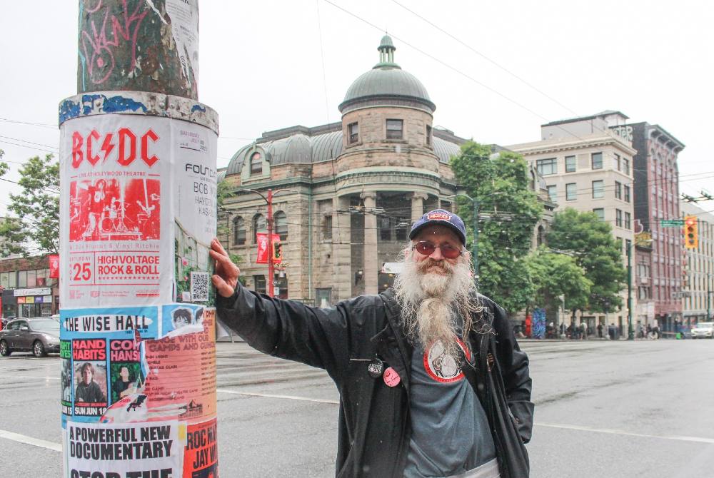 A 63-year-old man with a beard, hat and sunglasses leans against a lightpost at Main and Hastings in Vancouver. Behind him is a four-way intersection. There is an old classical building with a dome in the background.