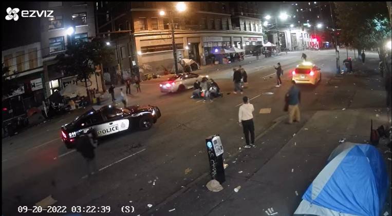 A screen capture of surveillance footage of a street at night. It is the aftermath of a car crash. A police car is parked in the middle of the road. A few people have clustered around a figure in front of the car, who is on the ground.