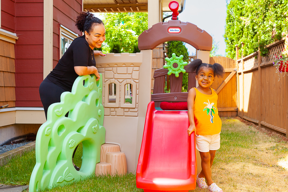 A mom and her kid stand beside a plastic kid’s playset.
