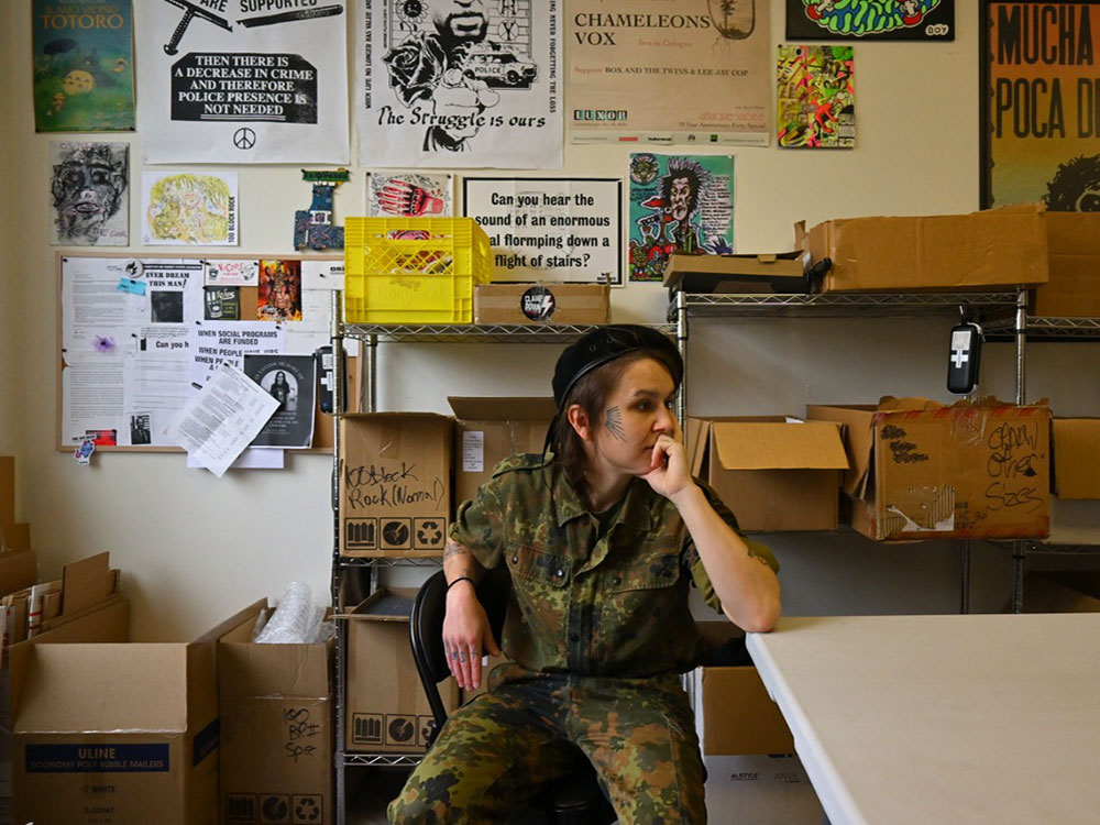 Nyx, a tattooed woman in camo, sits at a table in a room full of posters, and boxes of harm reduction supplies.